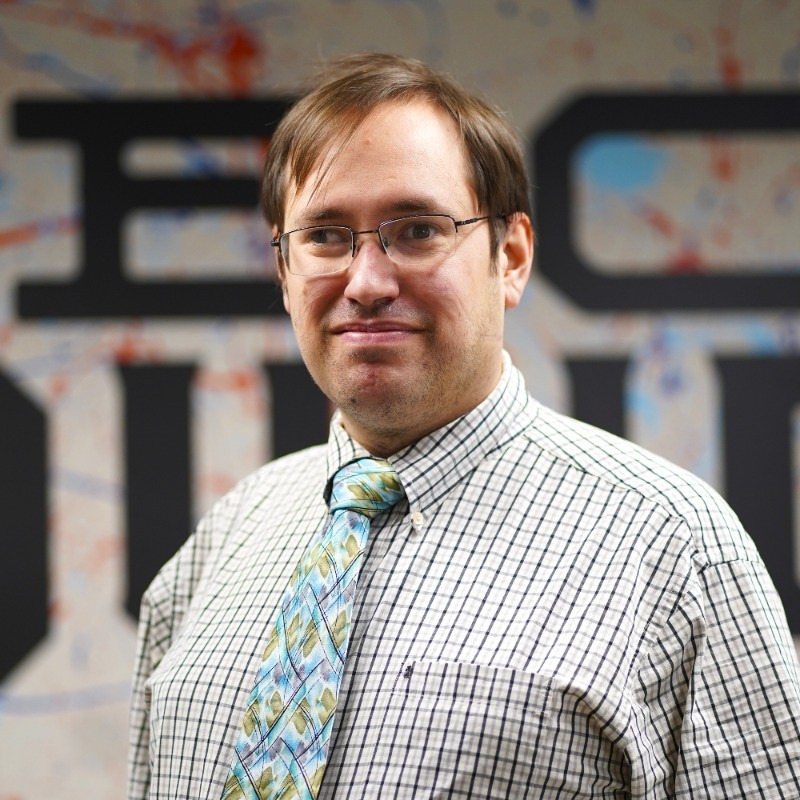 A man in a white shirt with a gray pattern and wearing a tie-dye patterned tie and glasses.